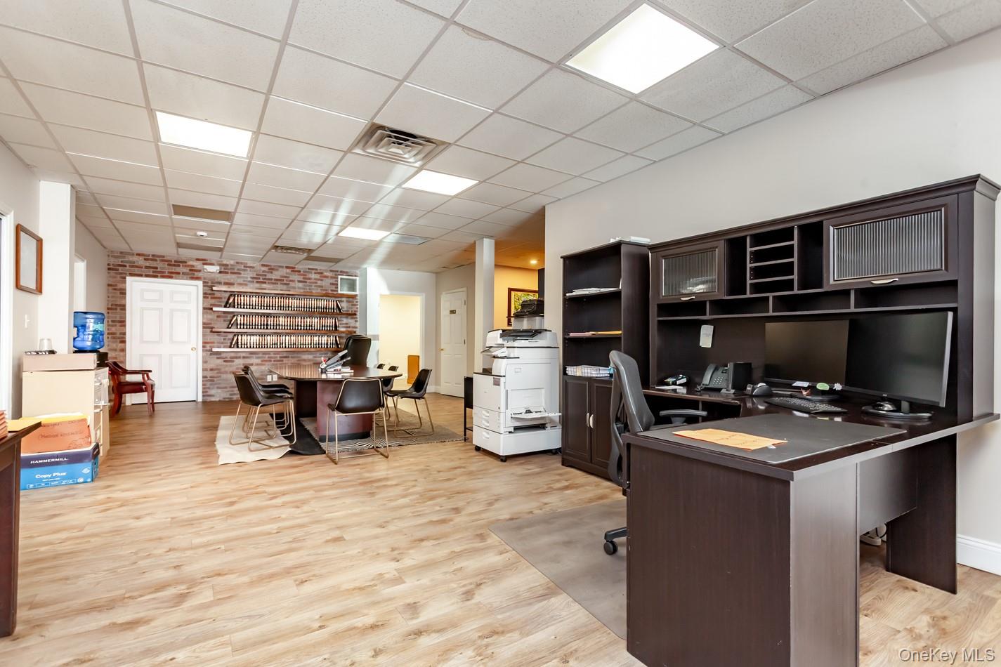 Office area with a drop ceiling, light wood-style flooring, and brick wall