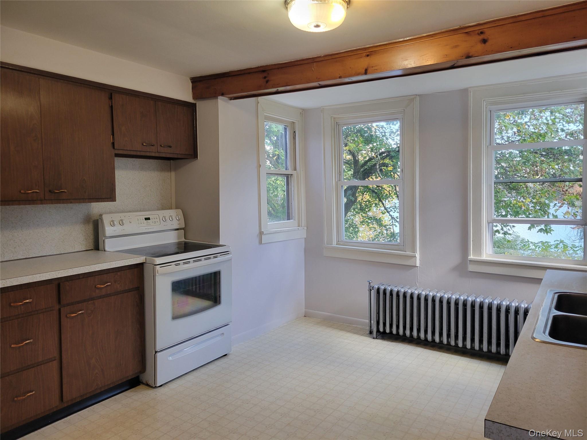 Kitchen with white electric range oven, dark brown cabinets, radiator heating unit, light countertops, and healthy amount of natural light