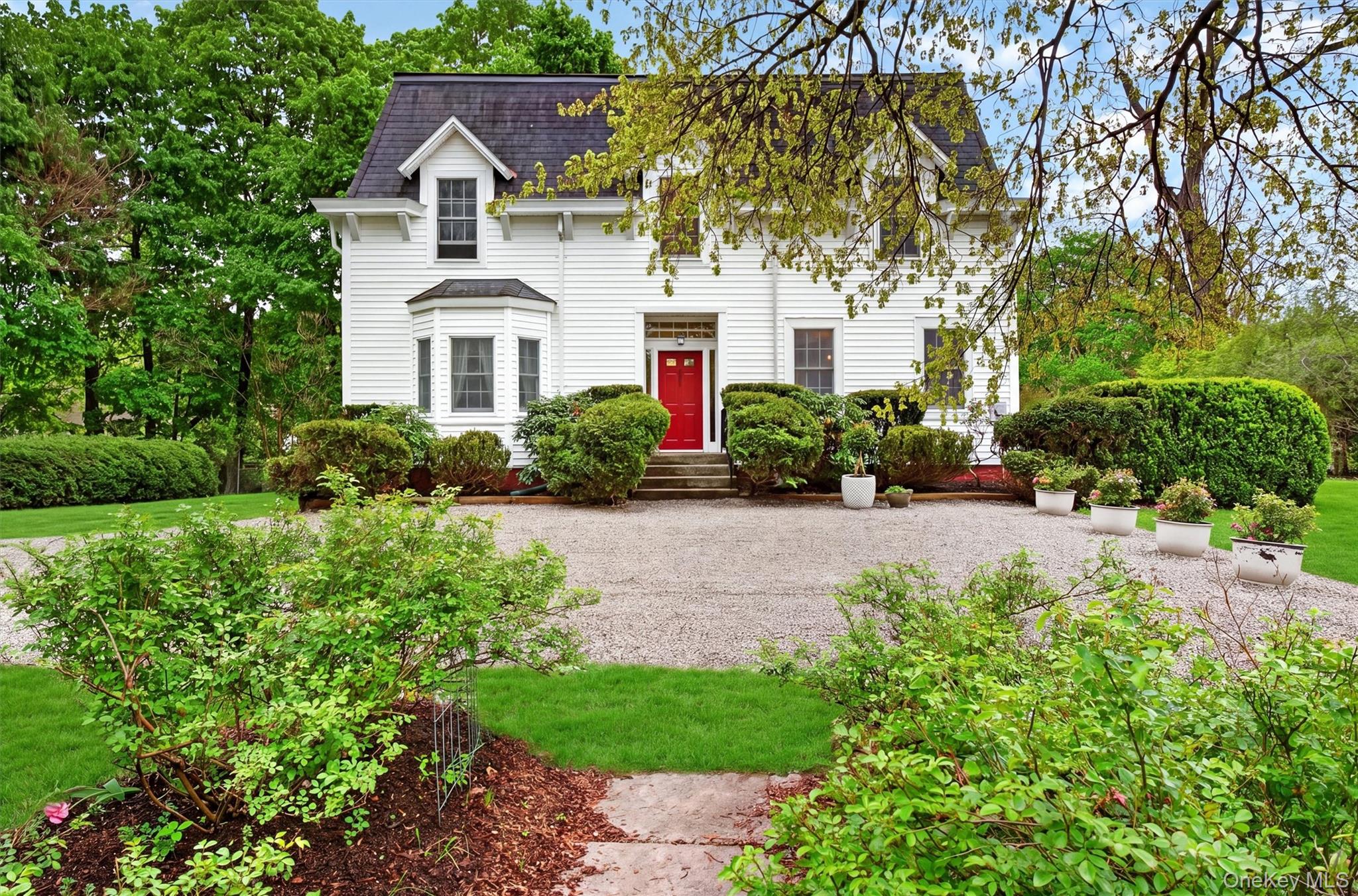 Exterior of colonial residence with red door and mansard roof. Gravel driveway with manicured gardens surrounding.