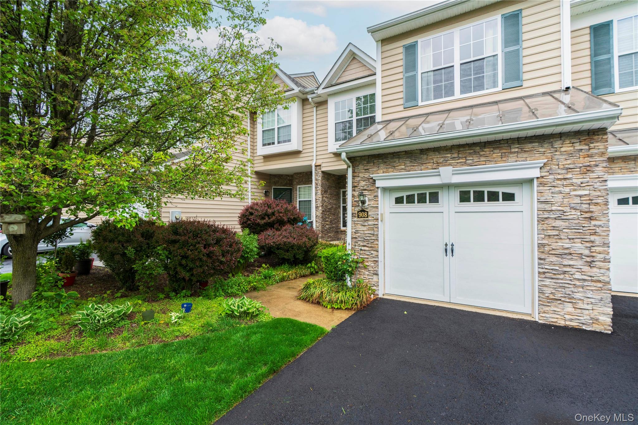 Exterior featuring stacked stone accents, vinyl siding, a two-car garage with paneling and windows, a glass canopy, and established landscaping
