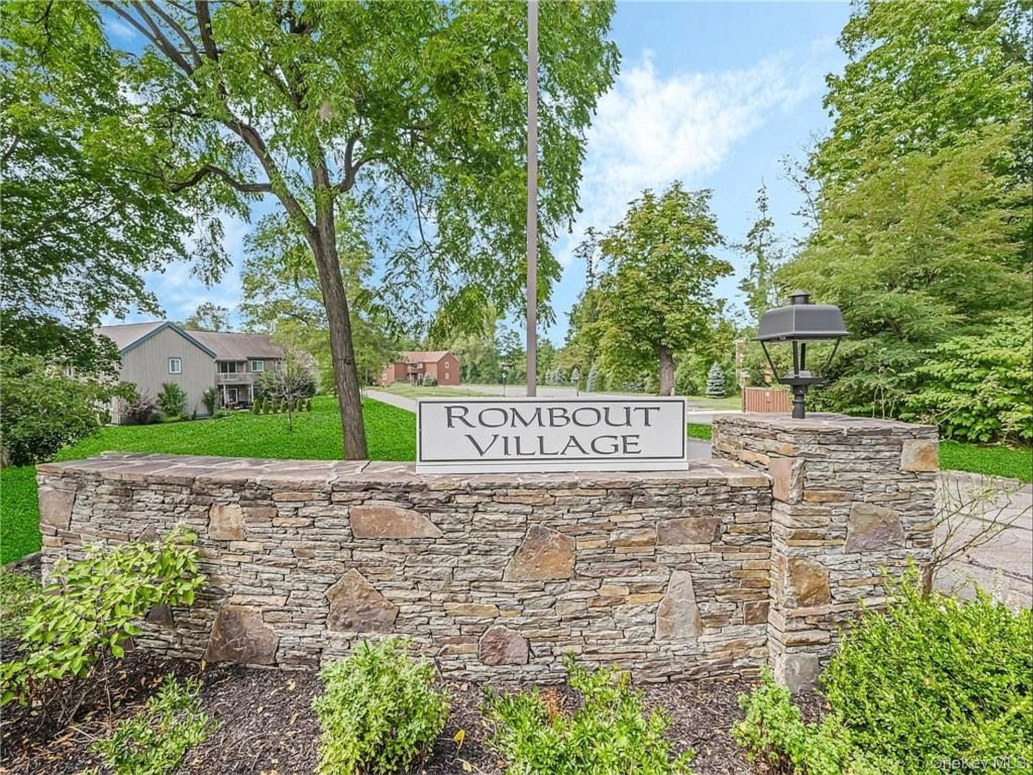 Stone-clad entrance wall featuring a prominent community sign, an integrated outdoor lamp post, and mature trees providing natural shading