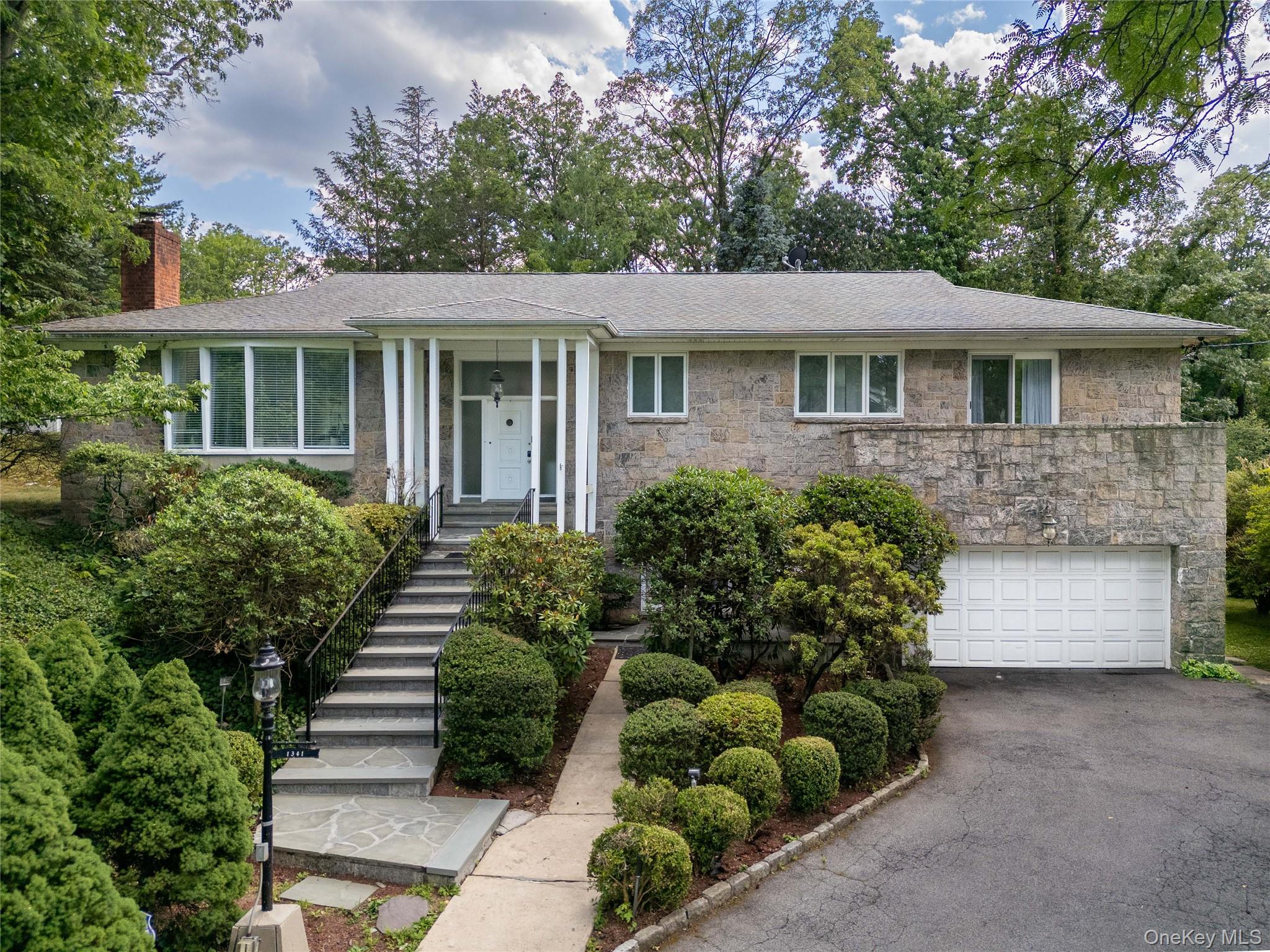 Stone facade residence featuring a prominent front entrance with white columns, a two-car garage, and extensive mature landscaping.