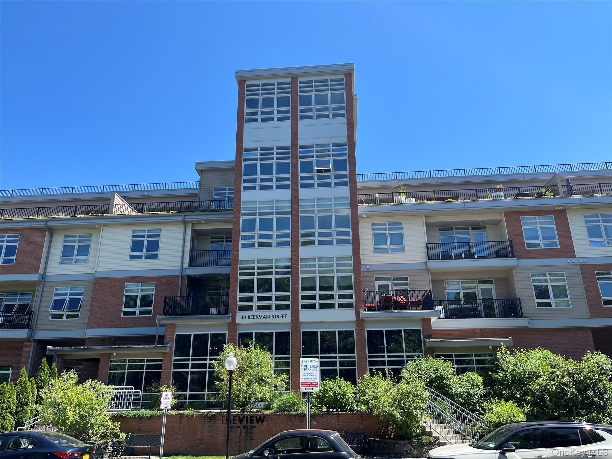 Multi-story residential building featuring a brick and siding exterior, expansive window panels, private balconies with black railings, and a prominent central glass-enclosed stairwell